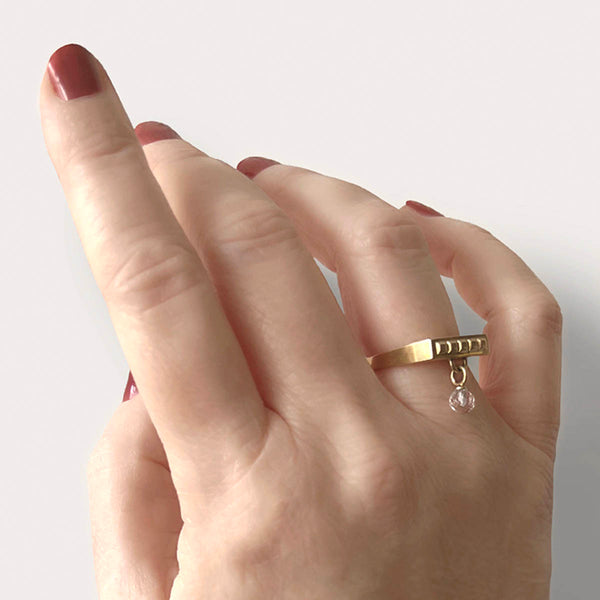 Square gold ring – Sleek square design with tiny studs and a clear quartz dangle charm on hand close up – Livva Østerby.