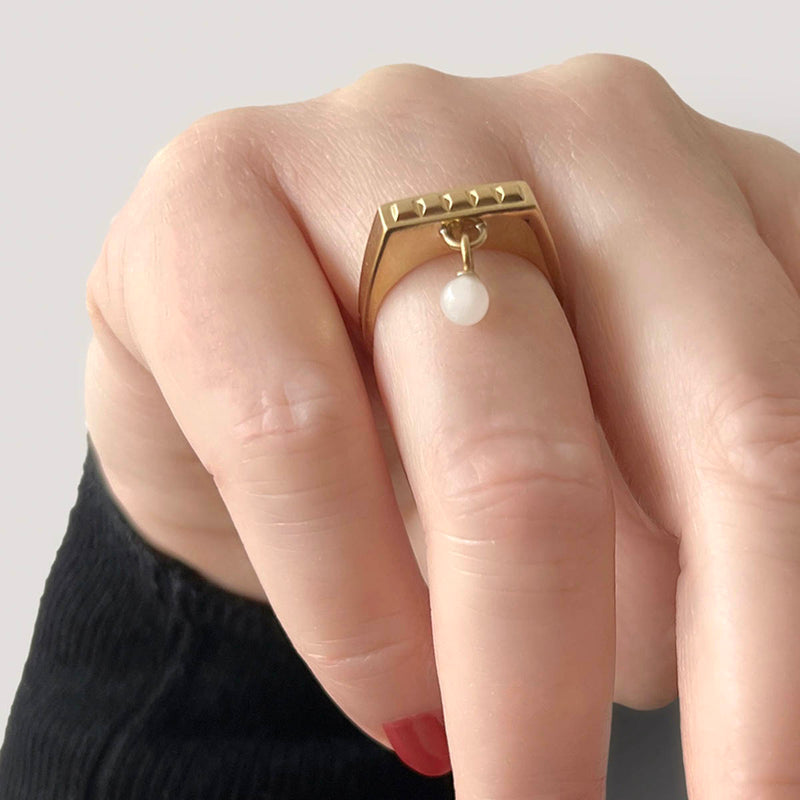 Square ring in gold – Sleek square design with tiny studs & a snow quartz dangle charm on hand close up – Livva Østerby.