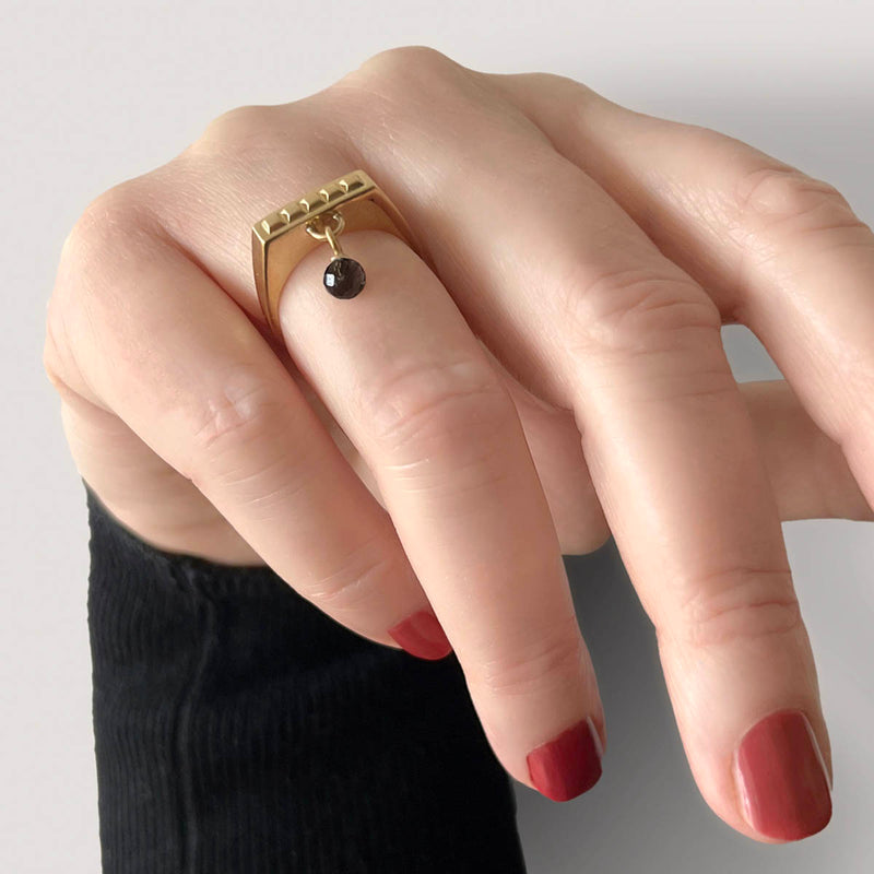 Square ring gold – A sleek square design with tiny studs and a black spinel dangle charm shown on hand – Livva Østerby.